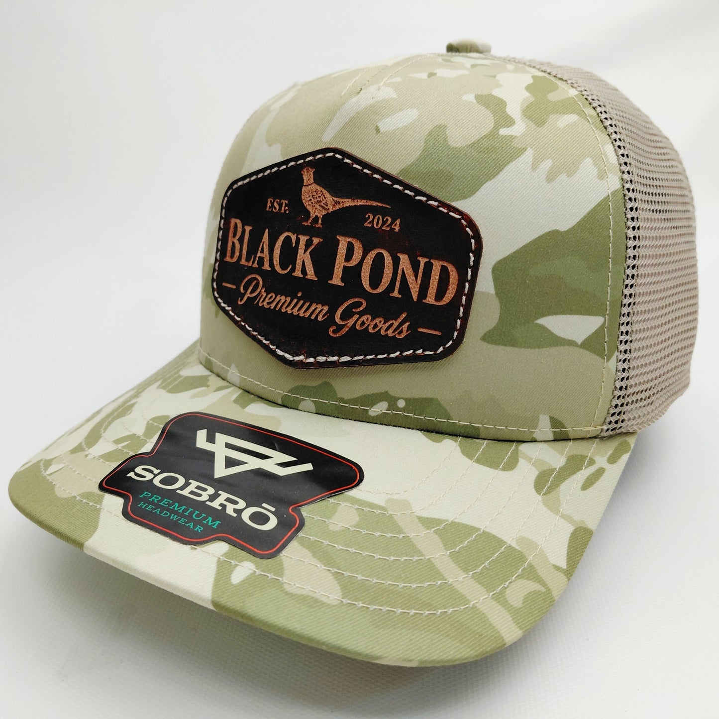 Left angled view of Black Pond pheasant leather patch SOBRO seven-panel tan camouflage trucker hat